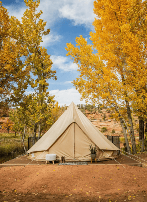 A beige tent surrounded by vibrant autumn trees under a blue sky, set on a wooden platform.